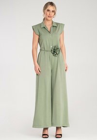Zielony kombinezon z krótkimi, wyściełanymi rękawami, kołnierzykiem, zapięciem na guziki, paskiem w talii i teksturalnym akcentem w formie kwiatu; wykonany z miękkiej, lekkiej tkaniny.