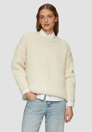 Dik gebreid crèmekleurig sweater met een ronde halslijn, ribbelstructuur en verlaagde schouders, gelaagd over een witte blouse met kraag.