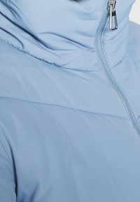 Světle modrá vyteplená bunda s vysokým límcem, která má prošívání a zapínání na zip. Hladká textura s matným povrchem.