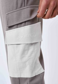 Grijze stoffen broek met textuur en een witte zakflap, versierd met de borduurtekst "Project x Paris". Zichtbare stiksels en een cargostijl.