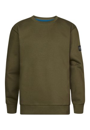 Person iført olivengrøn sweatshirt, blå jeans og hvide sneakers, stående mod en ensartet lys baggrund.