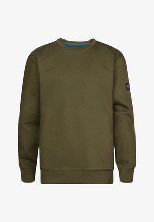 Personne portant un sweat-shirt vert olive, un jean bleu et des baskets blanches, se tenant devant un fond clair uni.