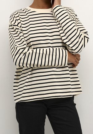 Osoba ubrana w białą koszulę z długim rękawem ozdobioną wąskimi czarnymi poziomymi paskami oraz czarne spodnie, z jedną złożoną ręką i drugą dotykającą twarzy.