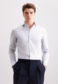 Giovane uomo con capelli corti castani che indossa una camicia a maniche corte a fantasia leggera e pantaloni plettati scuri, in piedi con le mani in tasca.