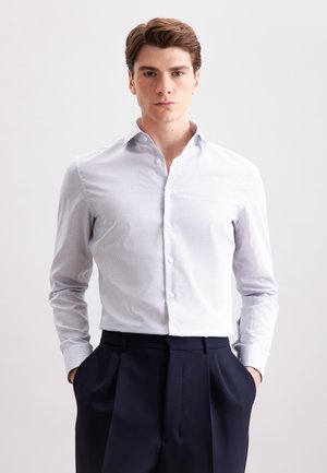 Giovane uomo con capelli corti castani che indossa una camicia a maniche corte a fantasia leggera e pantaloni plettati scuri, in piedi con le mani in tasca.