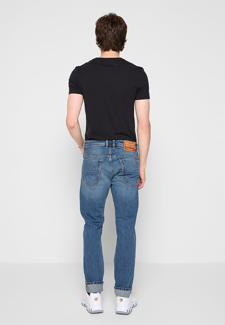 Homme portant un t-shirt noir, un jean bleu avec les ourlets repliés, et des baskets blanches, se tenant face à un fond gris clair uni.