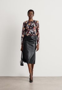 Bloemmotief blouse met lange mouwen in donkere tinten, gecombineerd met een zwarte leren rok tot de knie. Model houdt een kleine zwarte handtas vast.