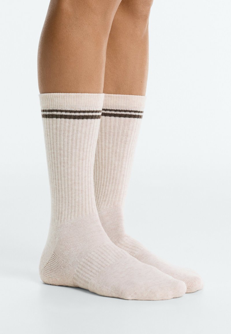 Jambes portant des chaussettes côtelées couleur crème mi-mollet avec deux bandes horizontales foncées près du haut sur fond blanc.