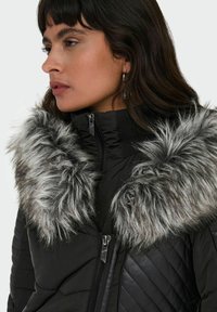 Veste noire matelassée avec un design matelassé, dotée d'un col en fausse fourrure grise détachable et d'une fermeture éclair. La texture semble lisse et douce.