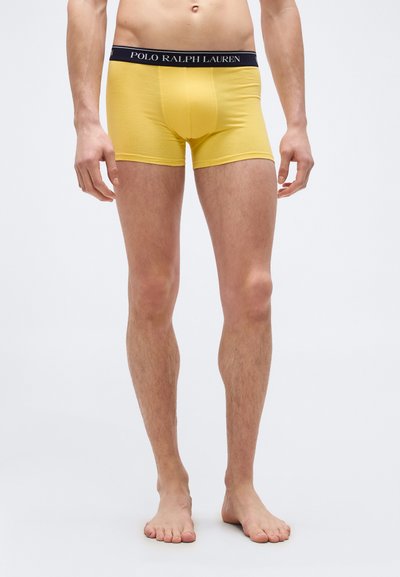 Άνδρας που φοράει κίτρινα μποξέρ με μαύρη μέση που γράφει «POLO RALPH LAUREN», στέκεται ξυπόλητος σε ανοιχτό γκρι φόντο.