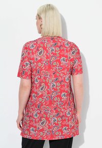 Červená krátkosrstá tunika s paisley a květinovými vzory. Hladká tkanina, volný střih a kulatý výstřih, sahající až k délce kyčlí.