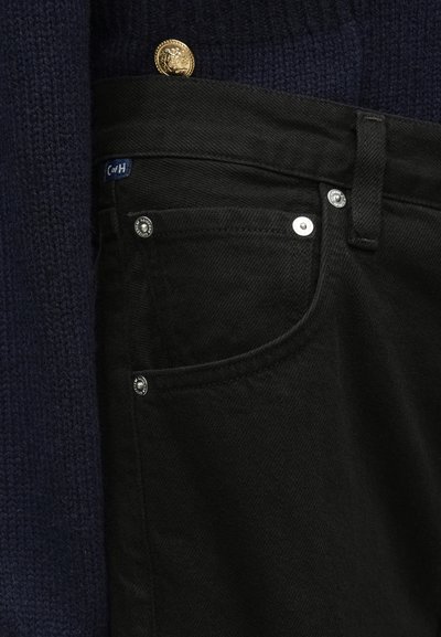 Čierne džínsové nohavice z denimu s tradičným dizajnom s piatimi vreckami, vybavené strieborným kovaním a modrou štítkovou značkou. Kombinované s tmavým pleteným svetrom.