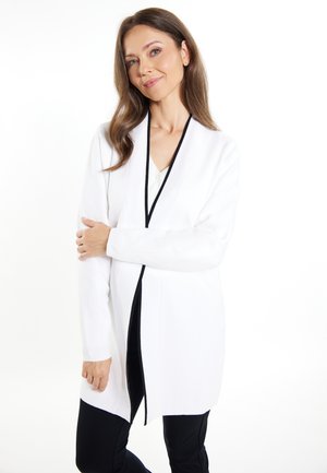 Cardigan lung alb cu dantelă neagră, cu față deschisă, textură moale și mâneci lungi. Asortat cu pantaloni închiși la culoare, evidențiind un croi relaxat.