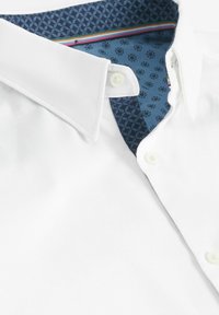 Camisa blanca con cuello puntiagudo, que muestra un forro interior de cuello azul con estampado y botones blancos en el frente.