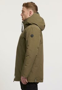 Olivgrüne isolierte Jacke mit Kapuze, Ansicht von rechts, mit einem runden schwarzen Logo am Ärmel und einem Reißverschluss am Saum.