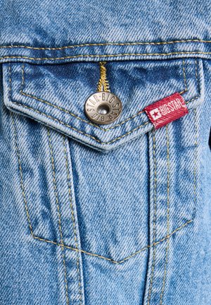 Svijetloplavi traper tkanina s oštrim džepom, srebrnim zakovicama i žutim šavovima, uz crvenu brendiranu oznaku sa strane.