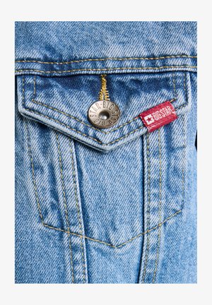 Gai zila džinsu audums ar smailu kabatu, sudraba poga un dzelteno šuvju, kā arī sarkanu zīmola etiķeti sānos.