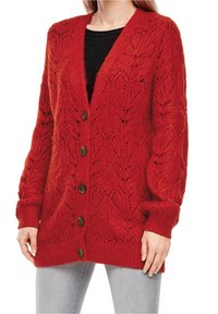 Femme portant un cardigan rouge en tricot avec des motifs en forme de cœur et de gros boutons, par-dessus un haut noir et un jean gris clair.