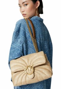 Borsa a mano intrecciata beige con un motivo testurizzato, un clasp ad anello dorato e una tracolla in catena dorata, indossata sopra una giacca di jeans.