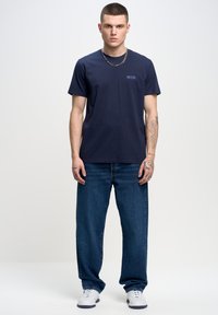 Jūras zils kokvilnas T-krekls ar īsām piedurknēm, ar mazu izšūtu logotipu kreisajā pusē. Saskaņots ar brīviem ziliem džinsiem un baltām kedām.