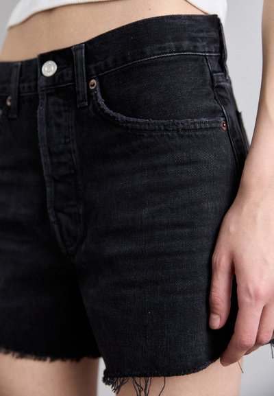 Detailná fotografia osoby v čiernych rifľových šortkách s ošúchanými lemy, bielom tričku a rukou pri boku.