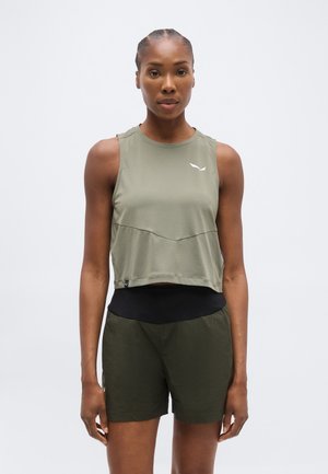 Kvinde iført ærmeløs olivengrøn træningstop og matchende shorts, stående med front mod kameraet mod en ensfarvet hvid baggrund.