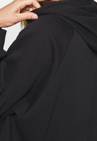 Sudadera con capucha negra de textura suave, que presenta un corte holgado y sutiles detalles de costura alrededor del cuello y los hombros.