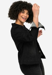 Veste en denim noir avec fermeture à boutons, poches latérales et accents en tissu gris sur les manches. Le modèle présente une vue de côté.