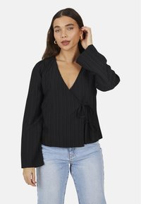 Top avvolgente nero con righe verticali, scollatura a V profonda, maniche lunghe, dettaglio legato in vita, abbinato a jeans azzurri chiari.