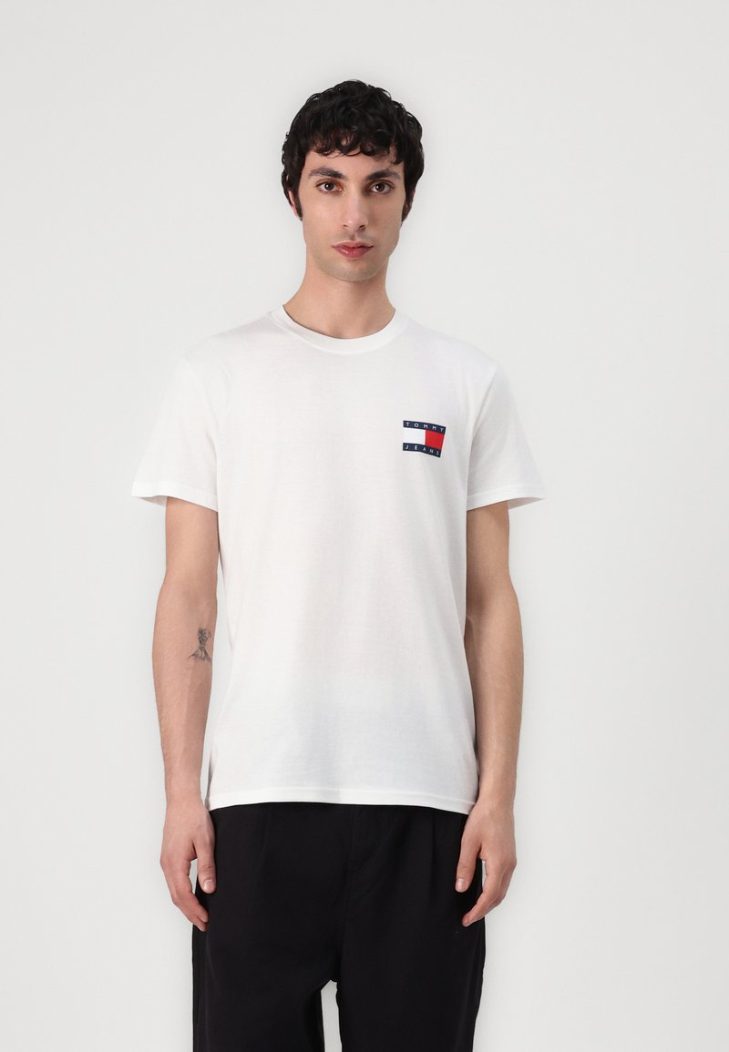 Camiseta blanca de algodón con un pequeño logo rectangular en azul marino y rojo en el lado izquierdo del pecho, cuello redondo y mangas cortas.