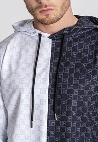 Pamučna majica s kapuljačom koja ima podijeljeni dizajn; jedna strana je bijela s uzorkom, dok je druga strana crna s sličnim uzorkom. Crne vezice.