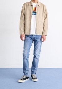 Giacca beige chiaro sopra una maglietta grafica bianca, abbinata a jeans azzurri e sneakers nere con suole bianche su un pavimento blu.