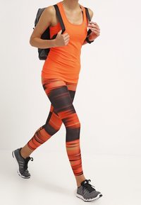 Débardeur de sport orange vif avec un legging noir et orange à motifs et des baskets grises. Les leggings présentent des rayures horizontales dynamiques.