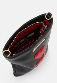 Schwarze Handtasche aus Kunstleder mit zwei roten Herzakzenten, goldfarbenen Beschlägen und einem Reißverschluss. Innenfutter aus rot gemustertem Stoff.