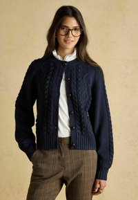 Tumši zila kapucīna džemperis ar adījuma raksta, ribotām piedurknēm, lielām brūnām pogām un garām piedurknēm, pārslāņots virs baltas apkakles krekla un svītrainām biksēm.