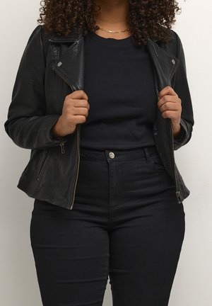 Person trägt schwarze Lederjacke, schwarzes Hemd, schwarze hoch taillierte Hose und goldene Kettenkette, hält die Revers der Jacke.