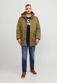 Άντρας φορεμένος με ελαιοπράσινο φούτερ με κουκούλα, ναυτικές μπλούζα με σχέδιο, μπλε τζιν και καφέ μπότες, στέκεται μπροστά σε ουδέτερο φόντο.