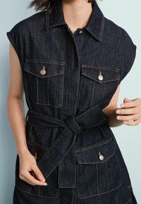 Denimowy kombinezon z krótkimi rękawami, wykonany z ciemnoniebieskiego materiału, z jasnobrązowym przeszyciem, dwoma kieszeniami na piersi i paskiem w talii. Wyposażony w srebrne zatrzaski.