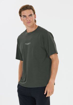 Jeune homme aux cheveux châtain clair portant un T-shirt à manches courtes vert foncé avec le texte "QUALITY ESSENTIALS" et un pantalon foncé sur fond blanc.