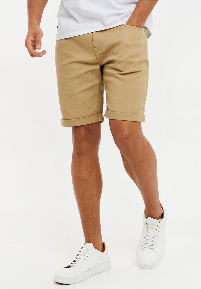Pantalones cortos caqui con dobladillos enrollados, hechos de una mezcla de algodón. Llevados con zapatillas blancas, que presentan una textura suave y un diseño con cordones.