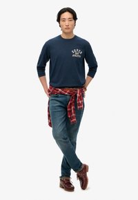 Camisa de manga larga de la marina con gráfico de "SUPER DRY ATHLETICS", jeans azules, camisa de cuadros roja atada a la cintura, zapatos marrones de estilo senderista.