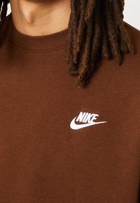 Brown cotton sweatshirt with a crew neckline. Features a white Nike logo embroidered on the chest. Fabric has a smooth texture.