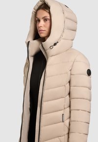 Bež puhasta jakna z velikim kapuco, s prešiti deli, z zadrgo spredaj in krožnim logotipom na levem rami.