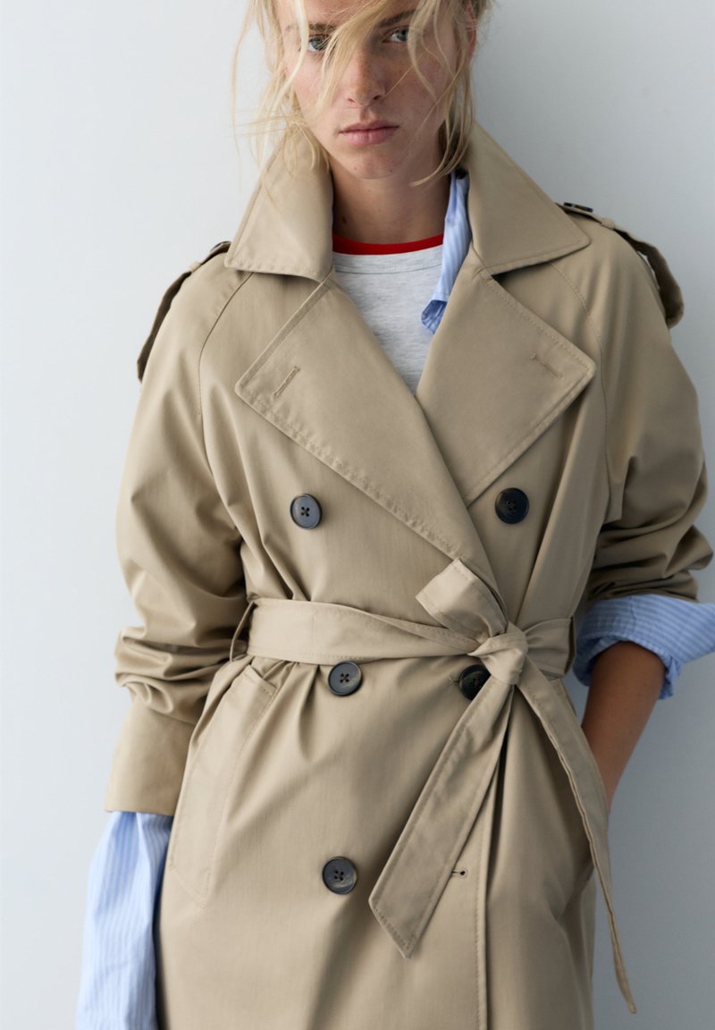 Lyshåret kvinde iført beige dobbeltradet trenchcoat bundet i taljen, lag på lag over blåstribet skjorte og hvid top med rød krave.