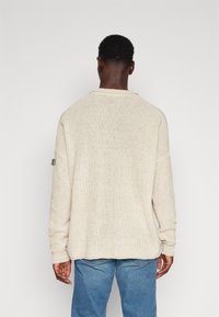 Helle beige Strickpullover mit lockerer Passform, weitem Kragen und gerippten Bündchen. Besitzt ein kleines Etikett an Ärmel links. Kombiniert mit blauen Jeans.
