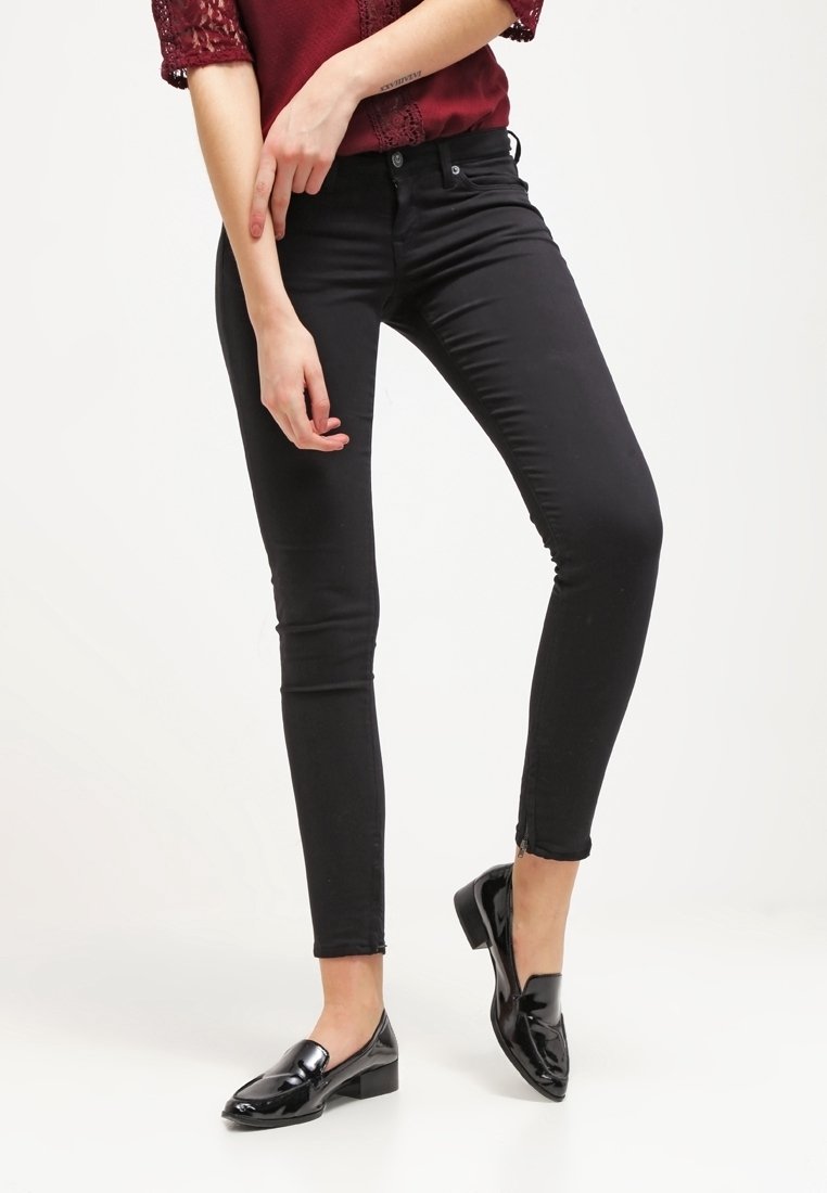 Kvinna som bär svarta skinny jeans, svarta lackläders loafers och en bordeauxfärgad blus med spetskanter, stående med benen i kors mot en vit bakgrund.