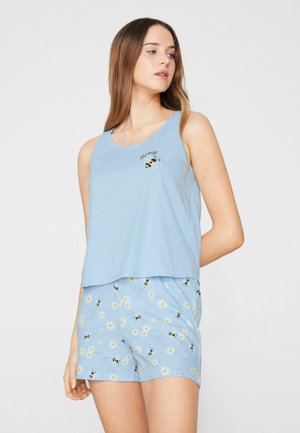 Femme portant un haut sans manches bleu clair avec un motif d'abeille et un short assorti avec un motif abeille et marguerite, debout sur fond blanc.
