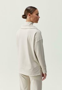 Moteris, turinti plaukus suktuke, vilki laisvą ilgomis rankovėmis smėlio spalvos palaidinę su aukštu apykakle ir derančias smėlio spalvos kelnes, stovi šone priešais vientisą foną.