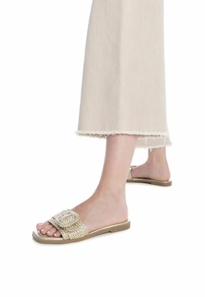 Sandali slide intrecciati in oro con una fibbia quadrata decorativa, suola piatta e soletta morbida, abbinati a pantaloni a gamba larga beige chiaro con orlo sfilacciato.