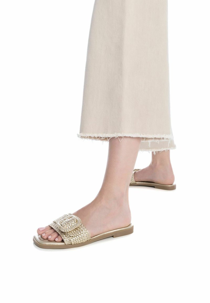 Sandali slide intrecciati in oro con una fibbia quadrata decorativa, suola piatta e soletta morbida, abbinati a pantaloni a gamba larga beige chiaro con orlo sfilacciato.